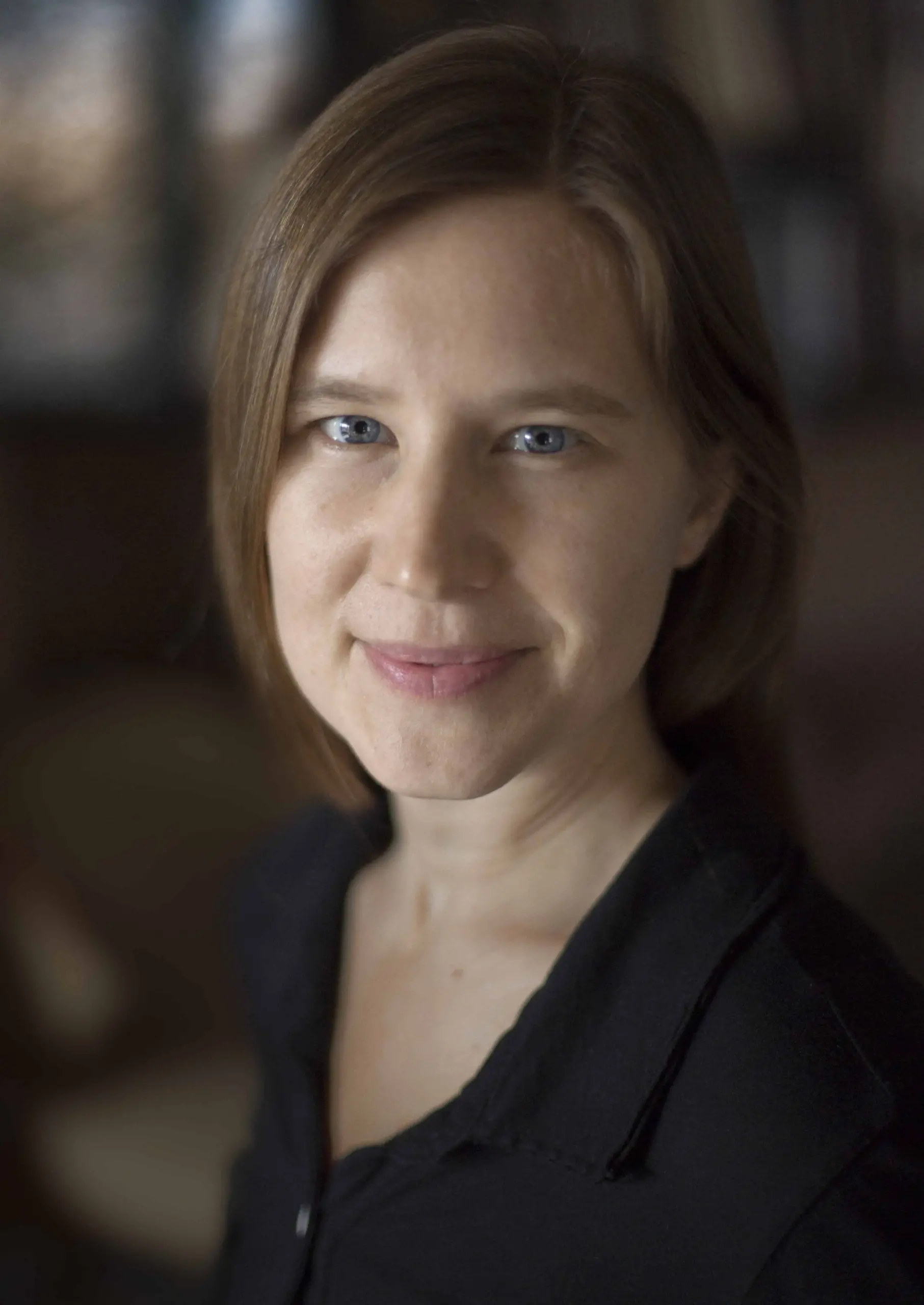 A woman with straight, light brown hair and blue eyes smiles softly at the camera. She is wearing a black top, and the background is softly blurred.