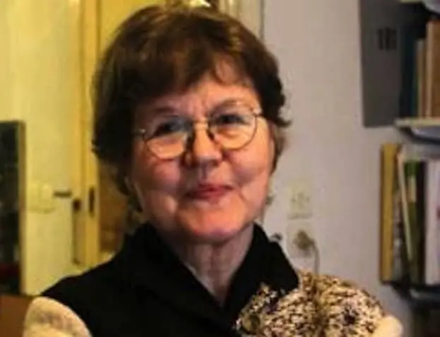 An older woman with short brown hair and glasses is smiling gently. She is wearing a dark top and standing indoors near a bookshelf. The background includes books and a light-colored wall.