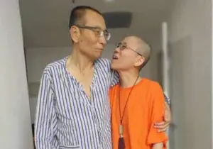 Two people with shaved heads stand closely together in a hallway. One wears striped pajamas, the other an orange shirt and glasses—reminiscent of Liu Xia’s emotive portraits—as they share an emotional moment, arms wrapped around each other.