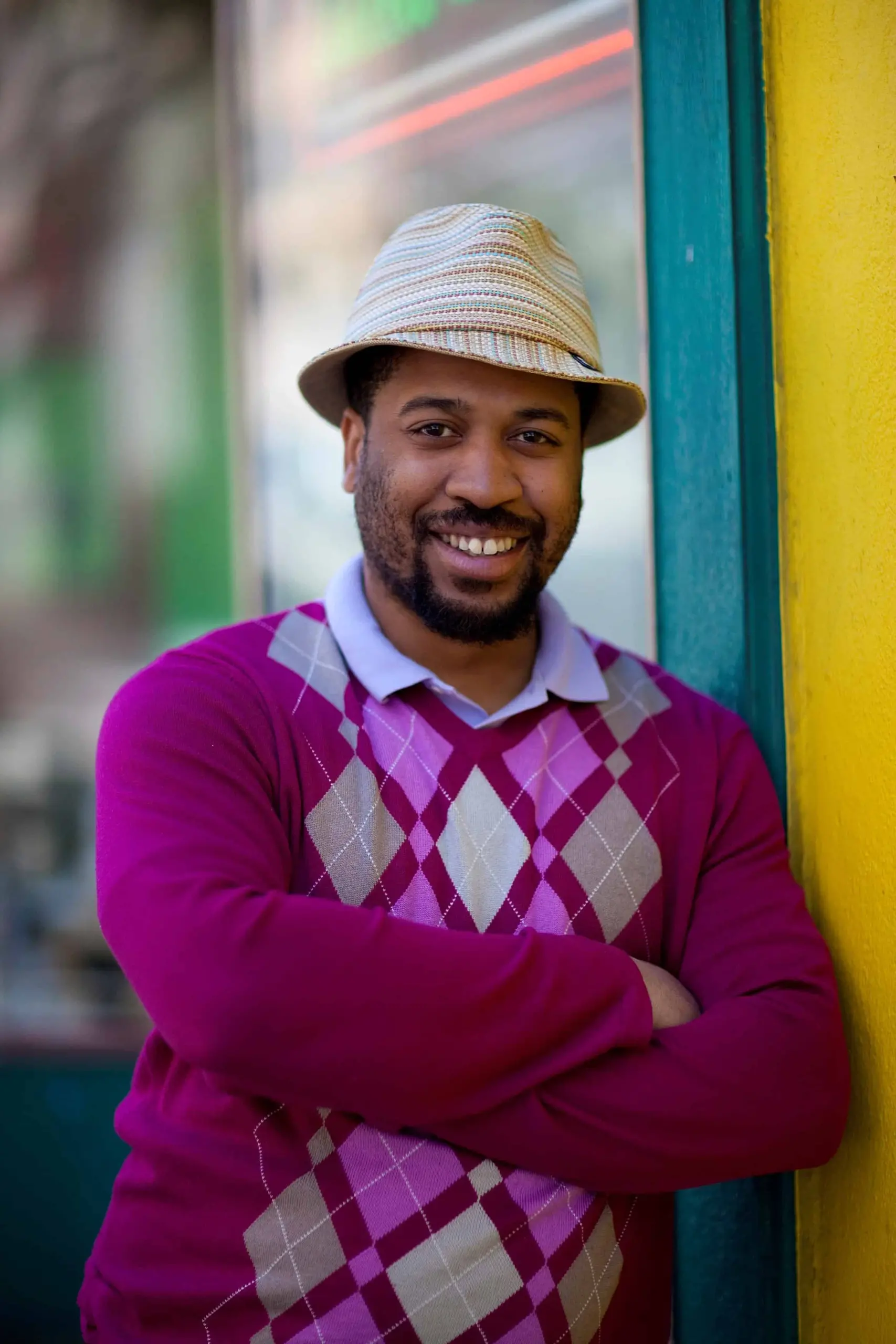A smiling man with a beard, wearing a beige hat and a purple argyle sweater, stands with his arms crossed against a yellow and teal wall.