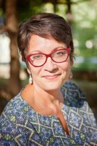 Meg Medina, a middle-aged woman with short dark hair and red glasses, smiles gently at the camera. She is wearing a patterned blue and green top and earrings, with a blurred outdoor background of trees and sunlight.