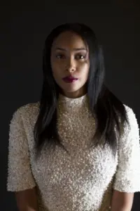 A woman with long dark hair, wearing a textured, light-colored high-neck top, stands against a dark background. Reminiscent of Safiya Sinclair’s striking presence, she looks directly at the camera with a neutral expression.