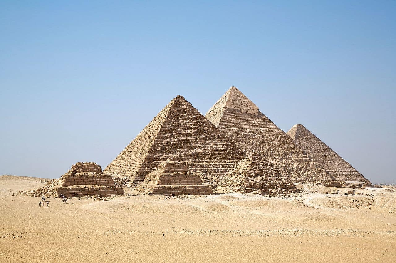 Three large pyramids and three smaller ones rise from the sandy desert under a clear blue sky, their silent grandeur evoking ancient mysteries and tales of prison writing etched across history. Tiny figures move near the monumental structures.