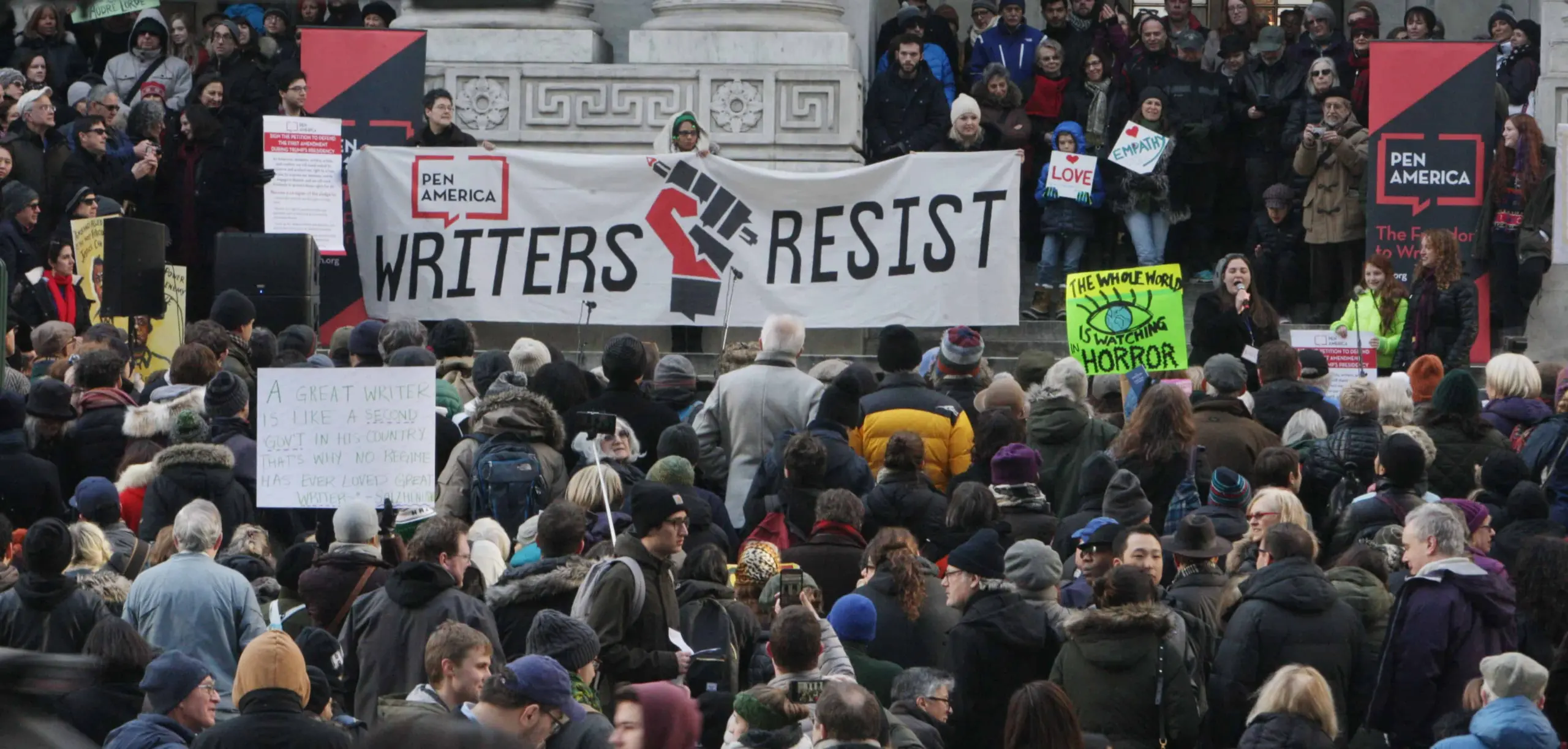 A large crowd gathers outdoors in winter clothing, holding signs including a large banner reading “Writers Resist” and a PEN America logo, at a protest in front of a stone building.
