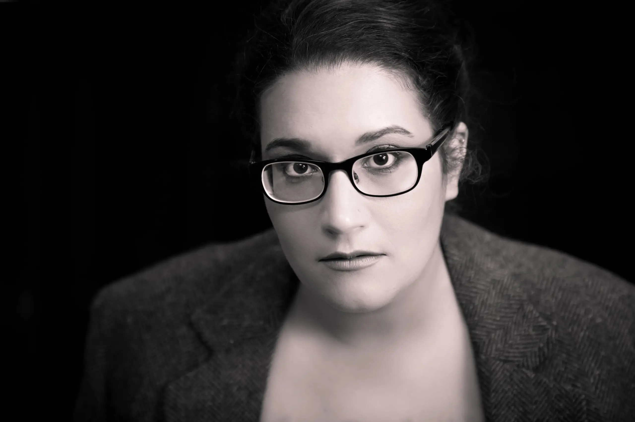A person with long dark hair pulled back, wearing glasses and a blazer, looks directly at the camera with a neutral expression—like an author captured in a classic literature portrait. The image is in black and white with a dark background.