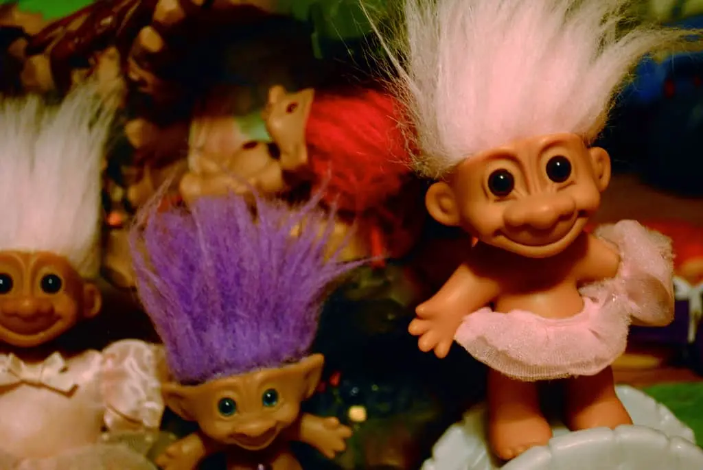 A group of vintage troll dolls with bright, wild hair in colors like white and purple, wearing small outfits and smiling, are closely gathered together.