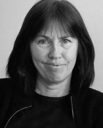 A woman with straight, shoulder-length dark hair and bangs is facing the camera, wearing a dark top and a slight smile. The image is in black and white.