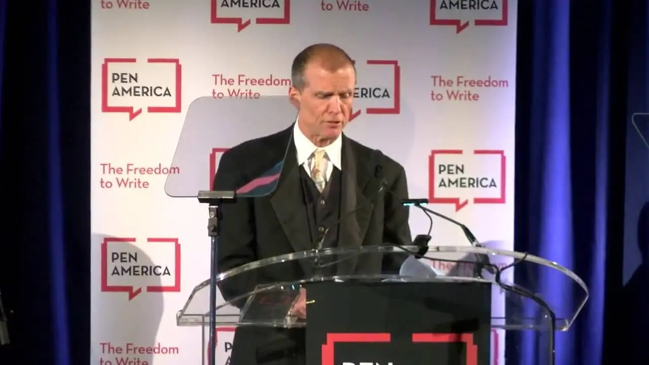 A man in a suit stands at a podium with a PEN America sign, speaking at an event. The backdrop displays repeating PEN America logos and the phrase The Freedom to Write.
