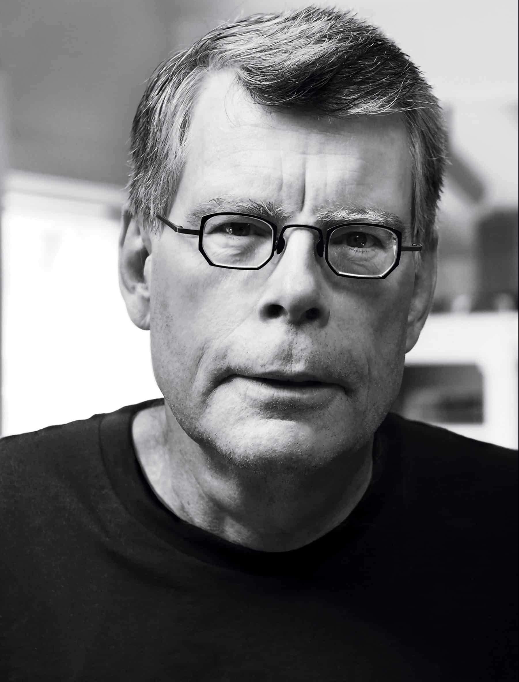 A man with short gray hair and glasses wears a black shirt and looks directly at the camera with a serious expression. The background is softly blurred in grayscale tones.