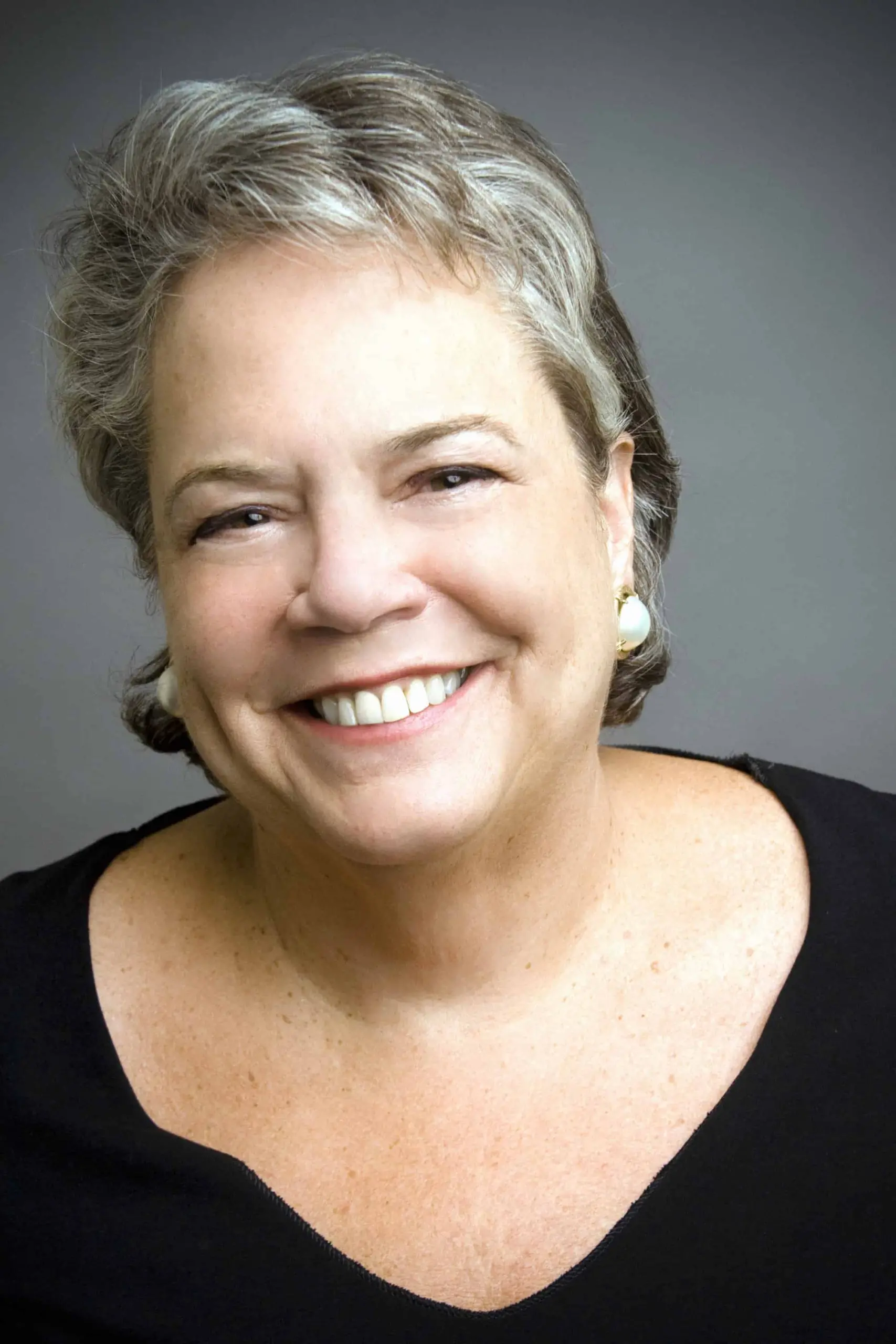 A woman with short gray hair and pearl earrings smiles warmly at the camera. She is wearing a black top and is set against a plain, softly lit background.