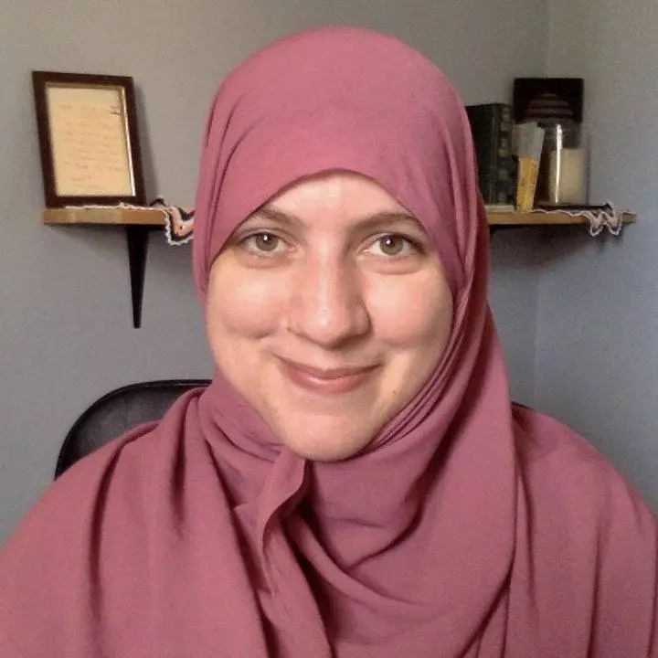 A person wearing a mauve hijab smiles at the camera. They are seated indoors with a shelf holding books, a framed picture, and a candle in the background.