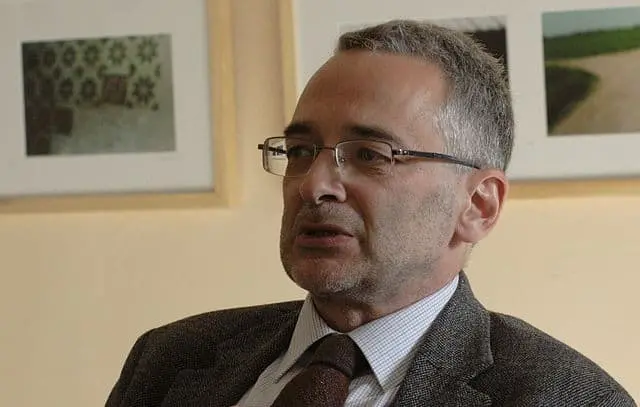 A middle-aged man with short gray hair and glasses, wearing a gray suit, striped shirt, and brown tie, sits indoors in front of framed photographs on a beige wall.