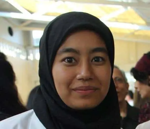 A woman wearing a black hijab smiles softly while standing indoors, with several people and bright lighting in the background.