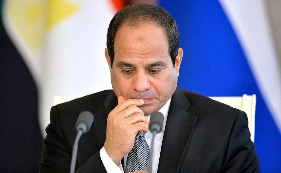 A man in a dark suit and tie sits at a table with microphones, appearing thoughtful, with his hand on his chin. Colorful blurred flags are visible in the background.