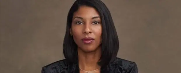 A woman with straight, shoulder-length black hair sits against a plain brown background, wearing a black jacket and layered necklaces, looking directly at the camera with a neutral expression.