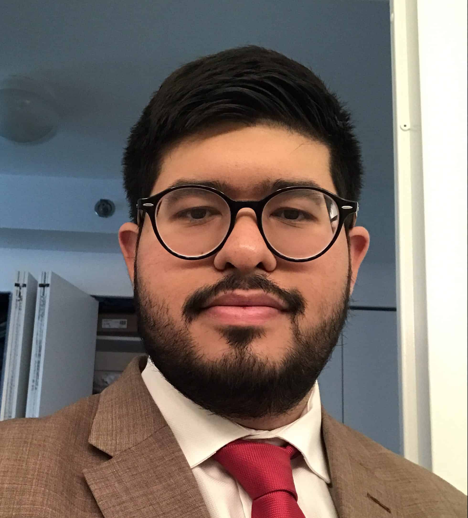 A man with short dark hair, a beard, and glasses, wearing a brown suit, white shirt, and red tie, stands indoors in front of an open closet door, smiling slightly at the camera.