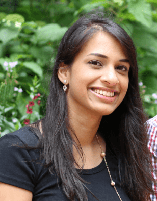 A woman with long dark hair and a black top smiles outdoors, with green foliage and purple flowers in the background.