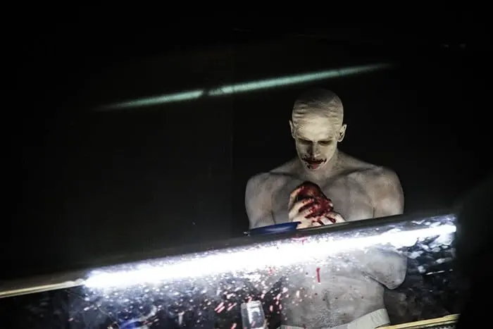 A shirtless person with pale skin and a bald head stands in the dark, gripping a red, heart-like object with both hands. Blood drips from the object, and their mouth is darkly stained, possibly with blood.