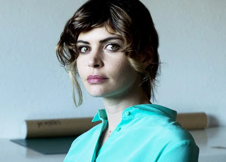 A woman with wavy brown hair and a side fringe, wearing a teal blouse, looks directly at the camera with a neutral expression. The background is plain with a tube resting on a surface behind her.