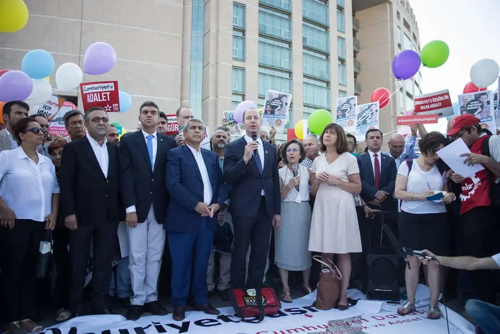 A group of people stands together at an outdoor rally holding signs and balloons. A man speaks into a microphone, while others, including men in suits and women, stand around him in front of a building.