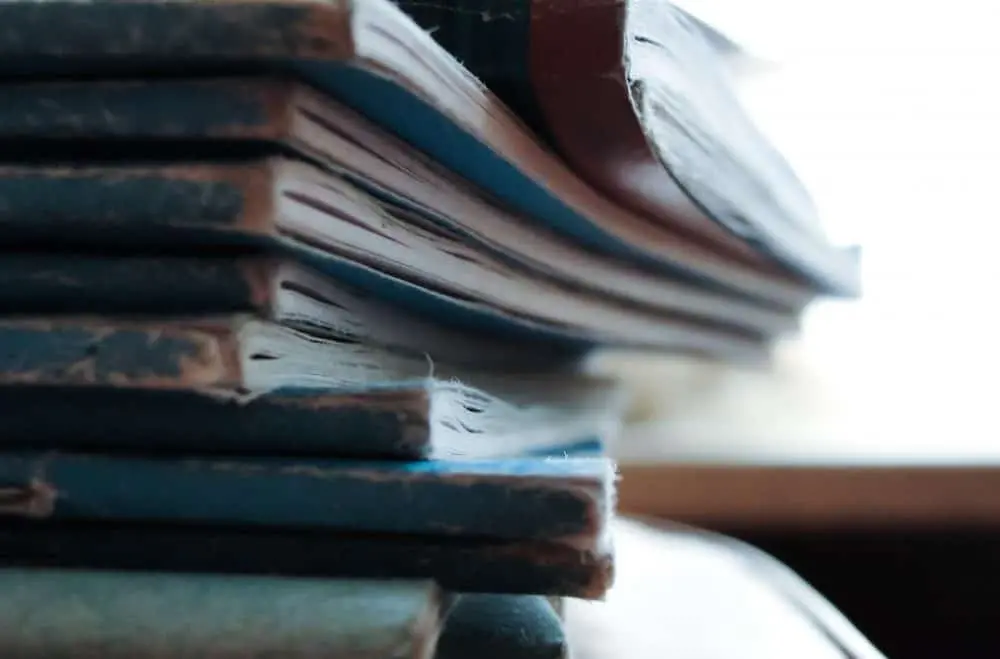 A close-up view of a stack of old, worn books with frayed edges and faded covers, arranged unevenly on top of each other. The background is softly blurred.