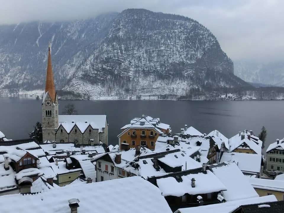 Snow-covered rooftops of a village with a tall church steeple, set beside a calm lake and backed by a large, tree-covered mountain partially shrouded in mist.