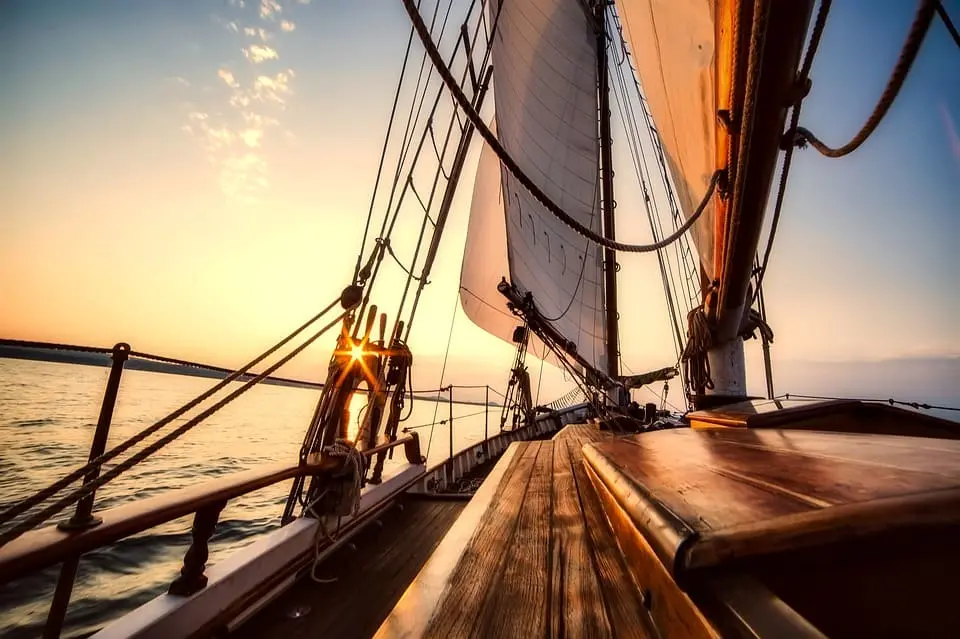 Sunset view from the deck of a sailing boat, with sunlight illuminating the sails, ropes, and wooden rails over calm water. The sky is partly cloudy with warm orange and yellow hues.