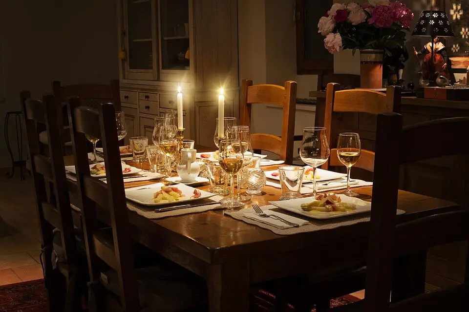 A wooden dining table set for six with plates of food, wine glasses, and two lit candles creates a warm, inviting atmosphere in a softly lit dining room. Flowers and decorative items are visible in the background.