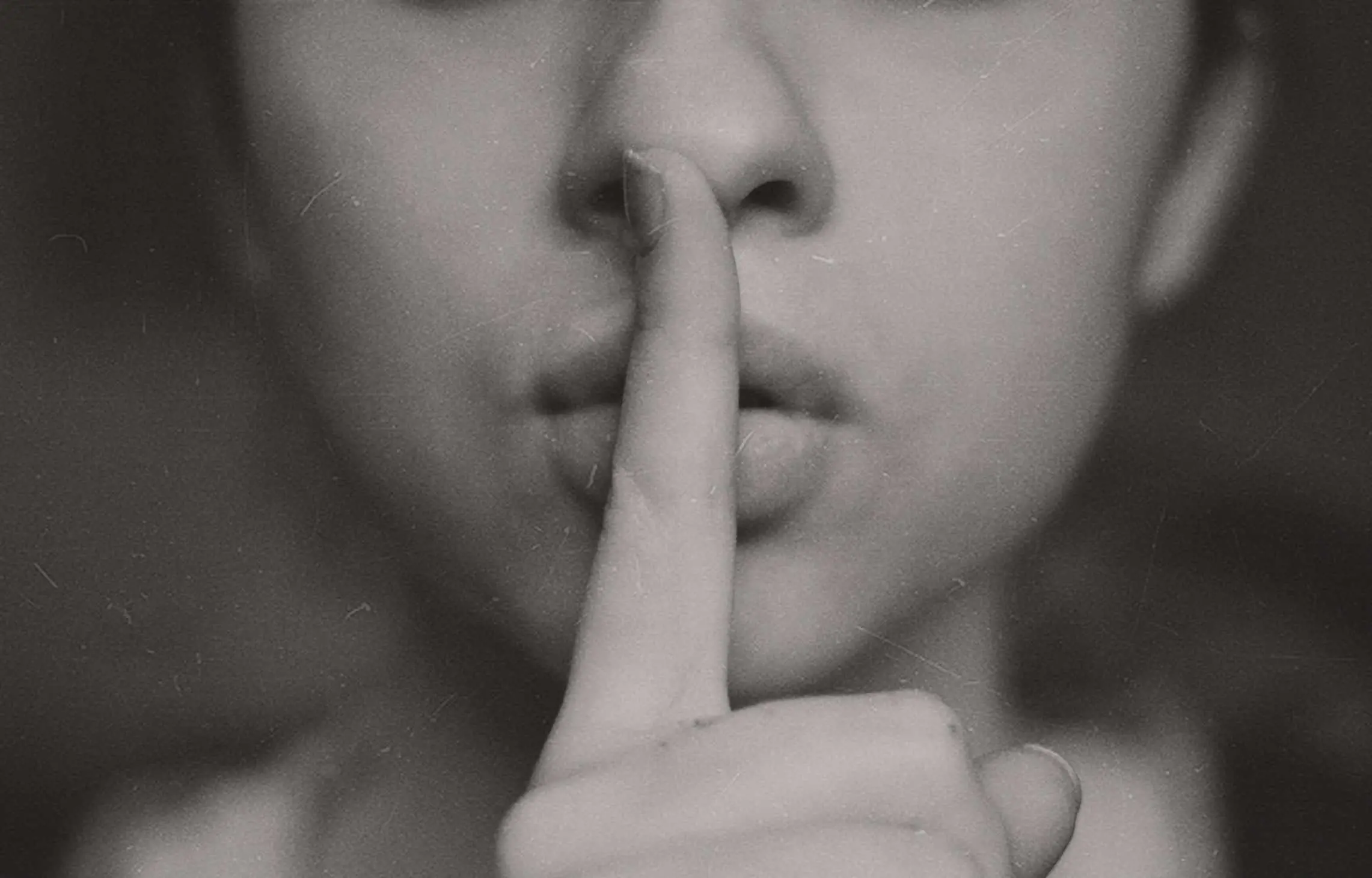 A close-up, black and white photo of a person holding a finger to their lips, signaling silence or a request to be quiet. The background is blurred, focusing on the lips and finger gesture.