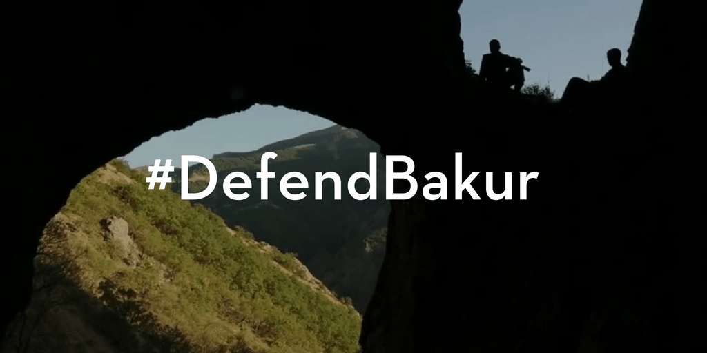 Silhouetted figures sit at the edge of a cave opening overlooking green hills and mountains, with the text #DefendBakur centered over the image.