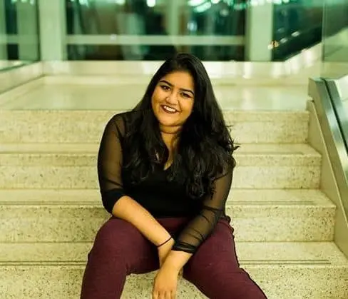 A woman with long dark hair, wearing a black top and maroon pants, sits smiling on a set of indoor stairs with glass railings on either side.