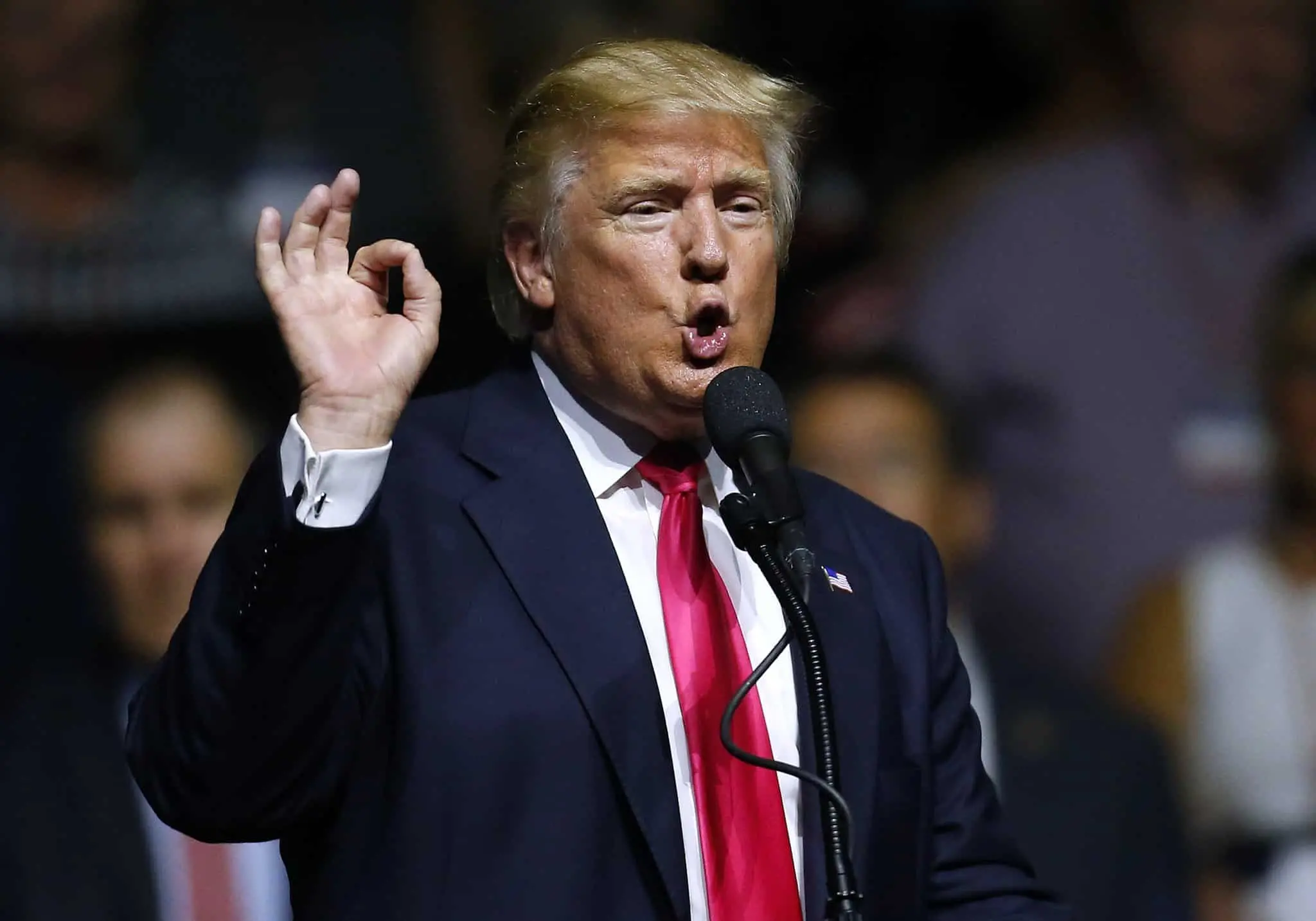 A man in a suit and red tie speaks into a microphone, gesturing with his right hand raised and making an OK sign. The background is blurred with people in shadow.