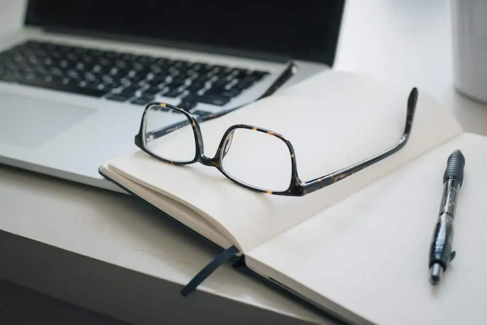 A pair of eyeglasses rests on an open notebook with blank pages, beside a pen, on a desk next to a laptop computer.