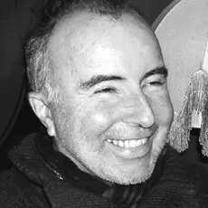 A middle-aged man with short hair and stubble smiles broadly. He is wearing a dark sweater, and the photo is in black and white. Part of a lampshade with fringe is visible in the background.