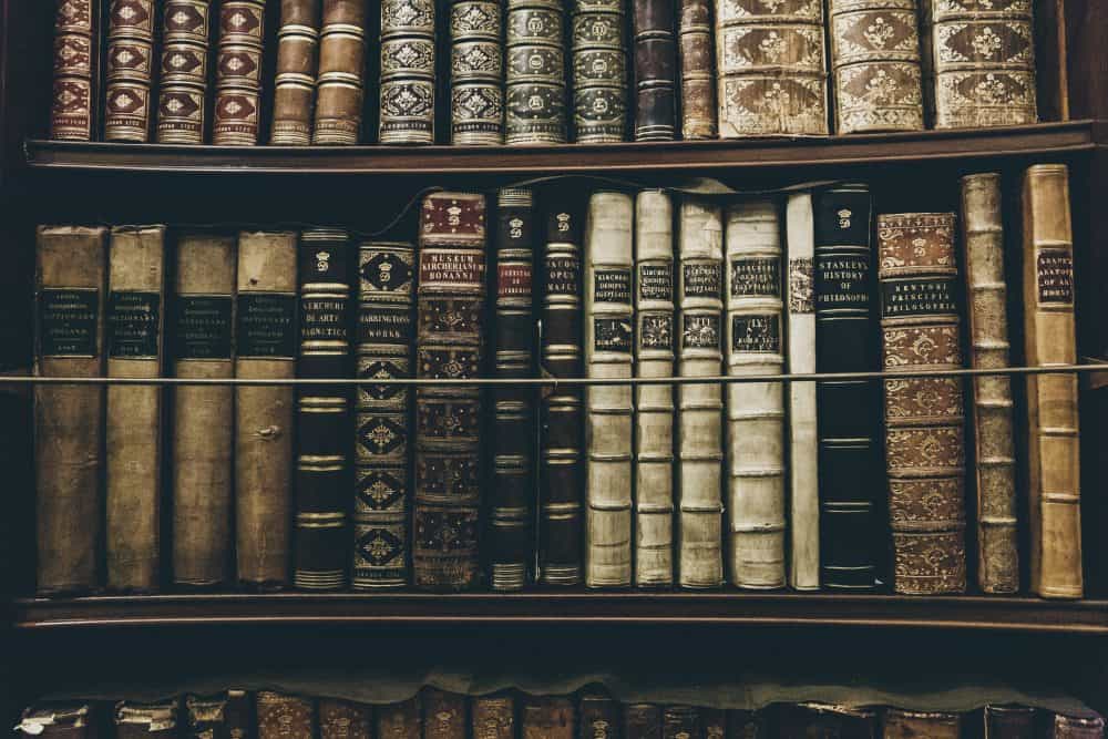 A close-up of shelves filled with old, antique books with ornate, worn spines in various shades of brown, beige, and black, showing intricate designs and faded text.