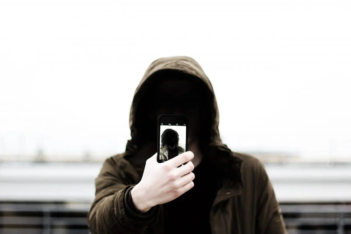 A person wearing a hooded jacket holds up a smartphone, taking a selfie. Their face is obscured by shadows, and the background is blurred and bright.