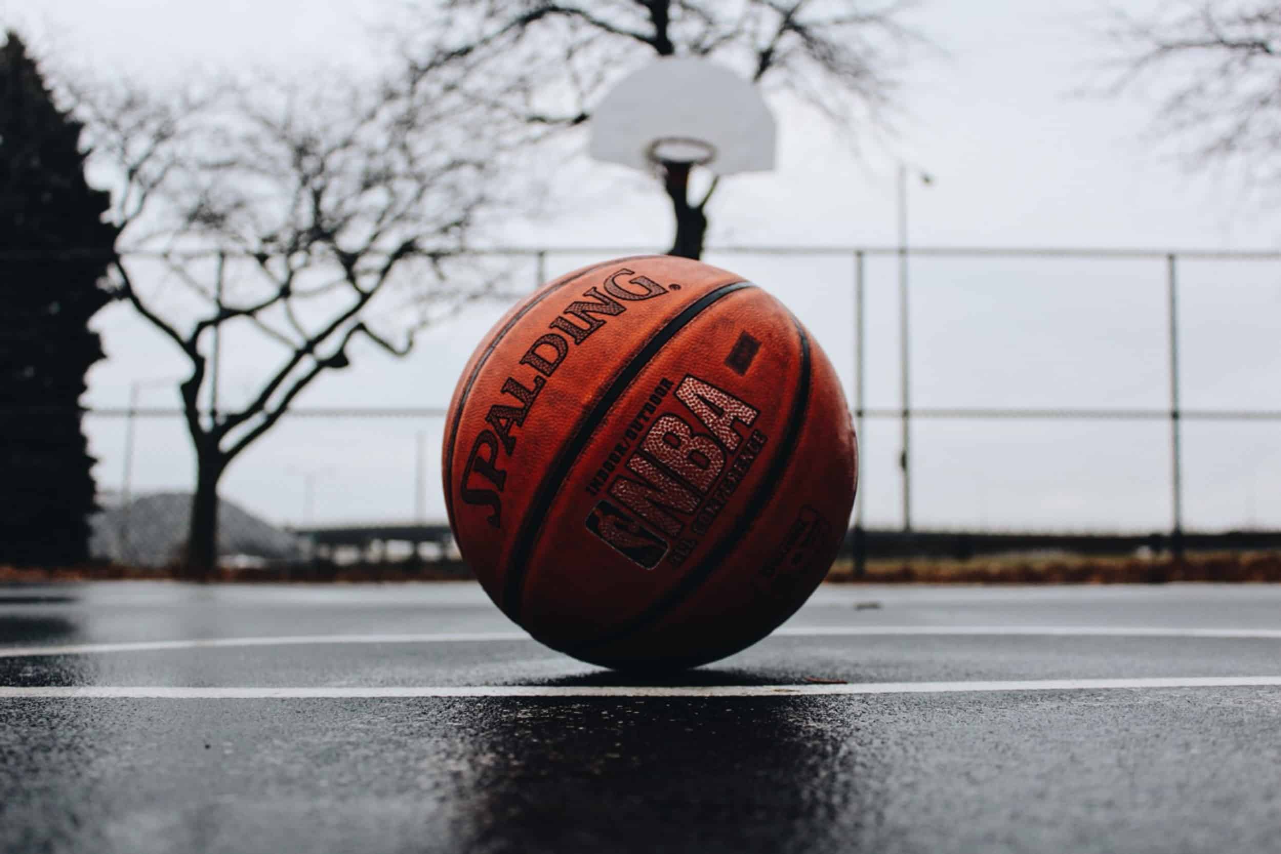 A Spalding NBA basketball rests on a wet outdoor court, with a hoop and leafless trees in the background on an overcast day.