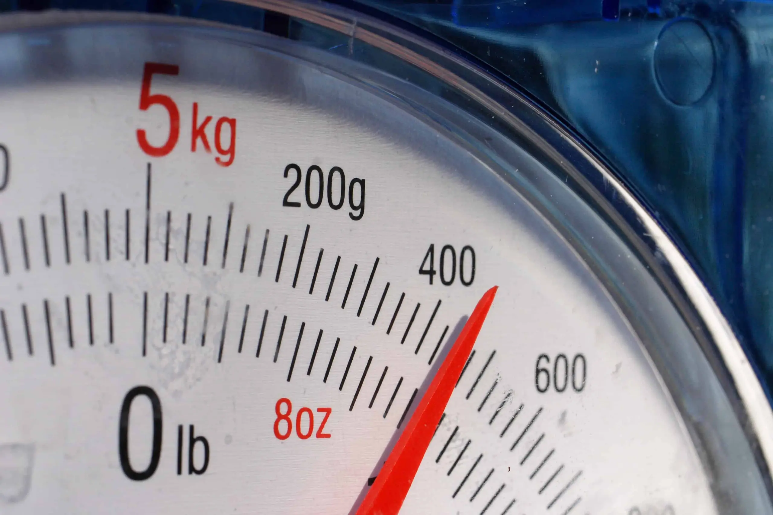 Close-up of an analog kitchen scale with a red needle pointing to approximately 400 grams. The scale displays measurements in grams, kilograms, ounces, and pounds.