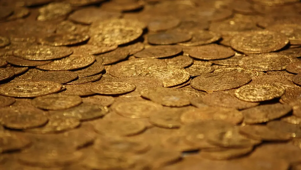 A close-up view of numerous shiny gold coins scattered and overlapping on a flat surface, creating a rich and textured golden background.