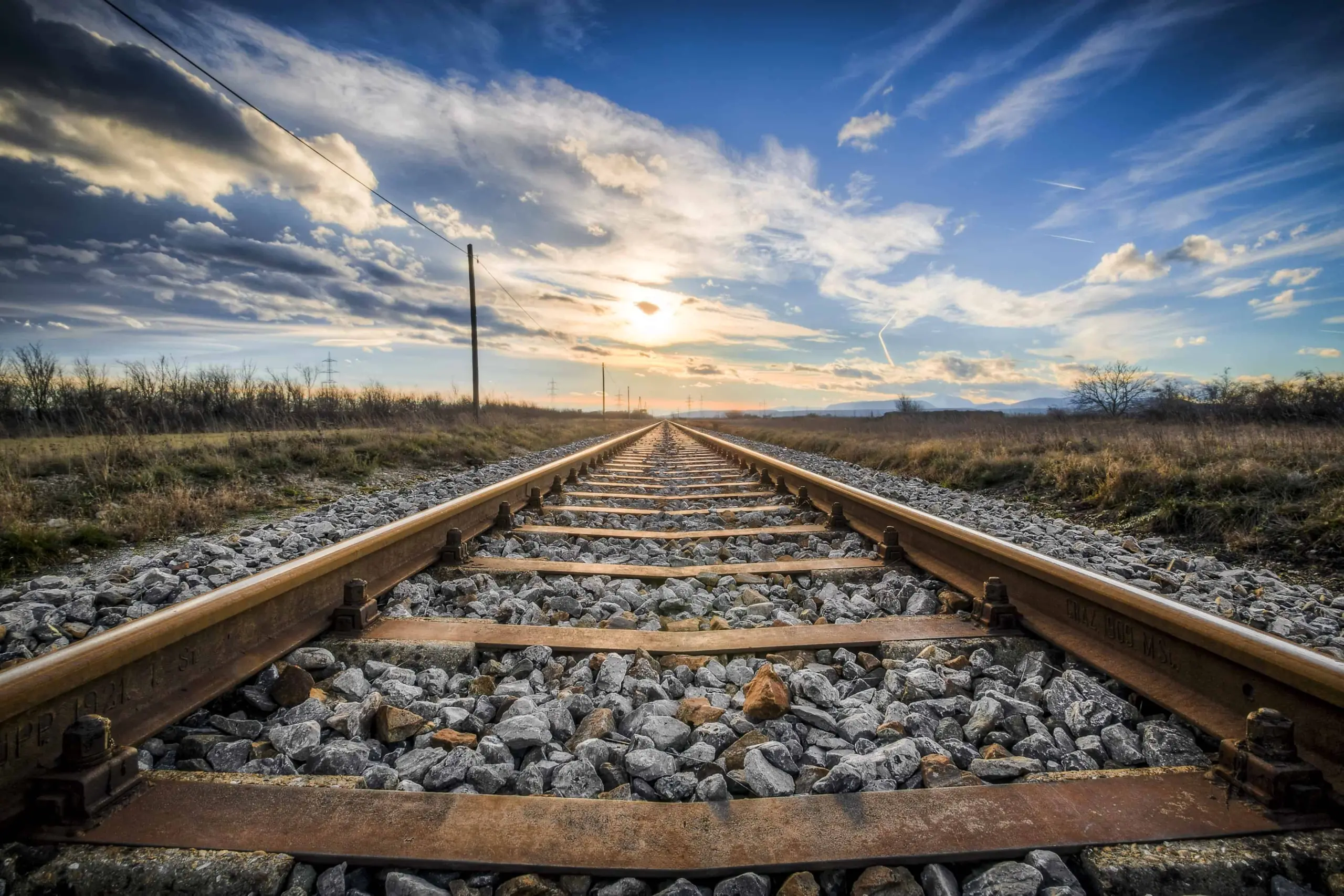 A set of railroad tracks stretches into the distance under a partly cloudy sky at sunset, with sunlight shining near the horizon and gravel covering the ground between and around the rails.