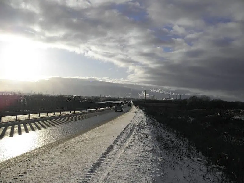 A snowy road stretches into the distance under a cloudy sky, with a few cars driving. The sun is low, casting long shadows and a bright glare on the road. Smoke rises in the background from industrial buildings.