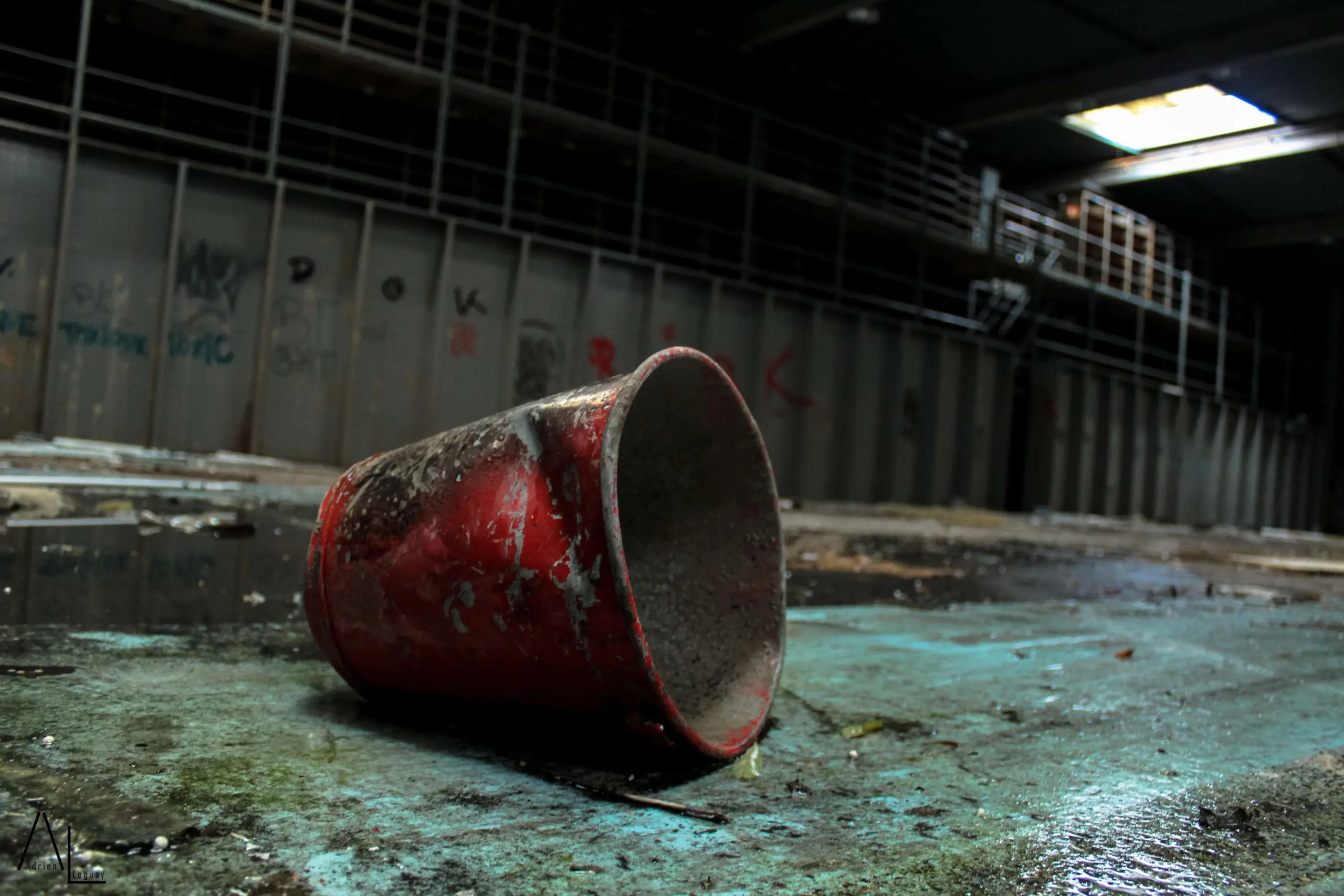 A dirty, red plastic cup lies on its side on a wet, blue-green floor inside an abandoned, graffiti-covered industrial building with dim lighting.