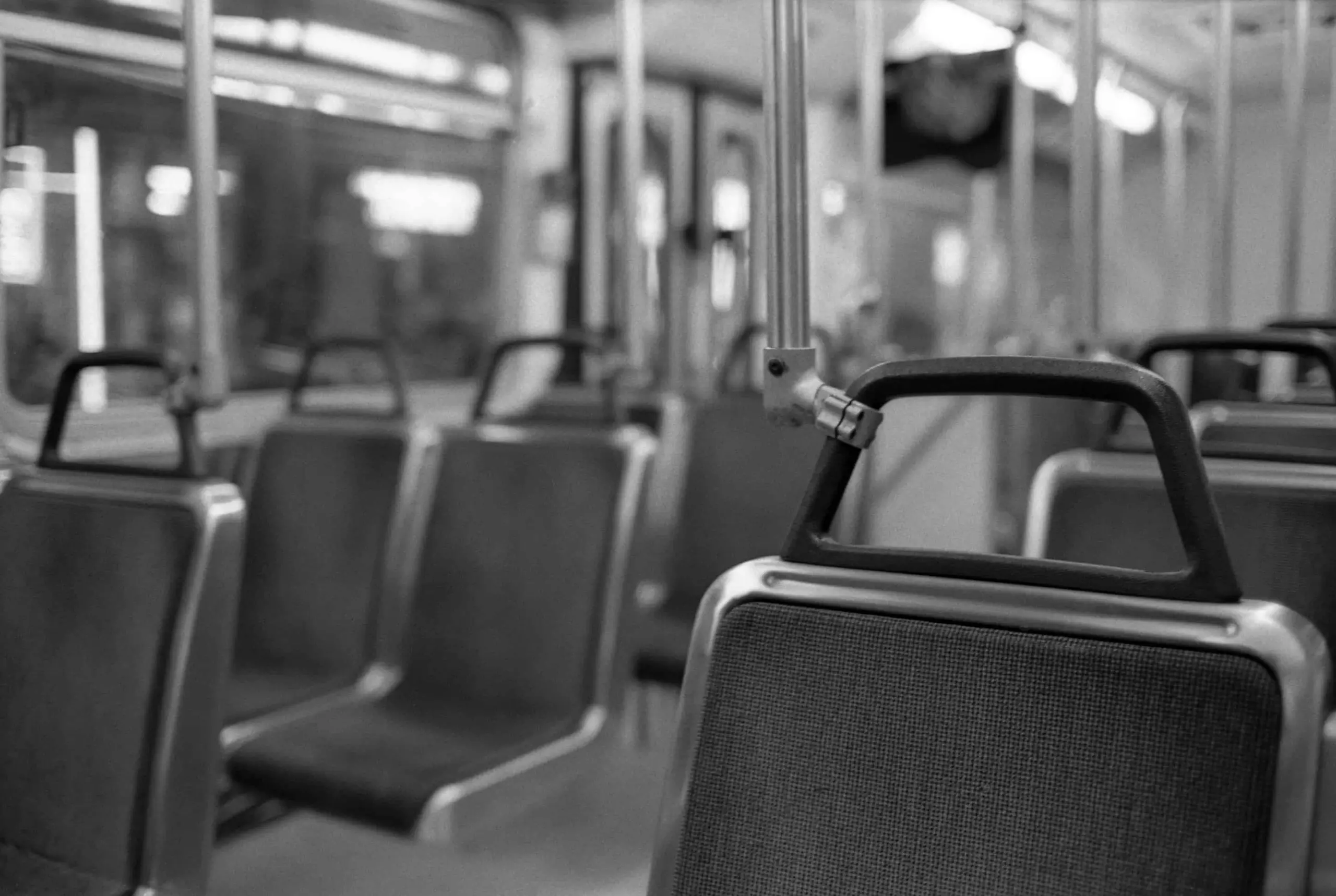 A black-and-white photo of empty seats inside a city bus or tram, with grab handles hanging from overhead bars and rows of unoccupied seats stretching into the background.