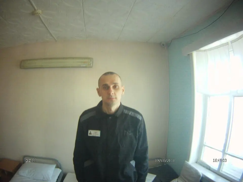 A man wearing a dark uniform stands in a simple, sparsely furnished room with light-colored walls, a bed, and a window letting in daylight. The photo appears to be taken with a wide-angle lens or security camera.