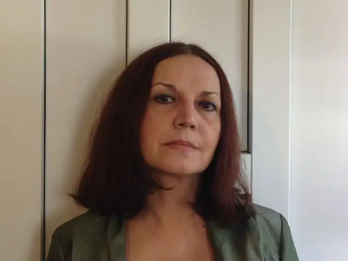 A woman with straight, shoulder-length dark hair and a neutral expression stands in front of a light-colored paneled wall, wearing a green top.