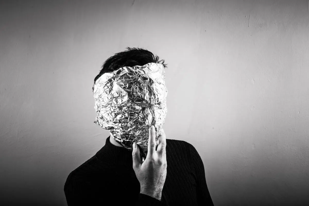 A person wearing a dark sweater holds a sheet of crumpled aluminum foil over their face, obscuring their features, against a plain light-colored background.