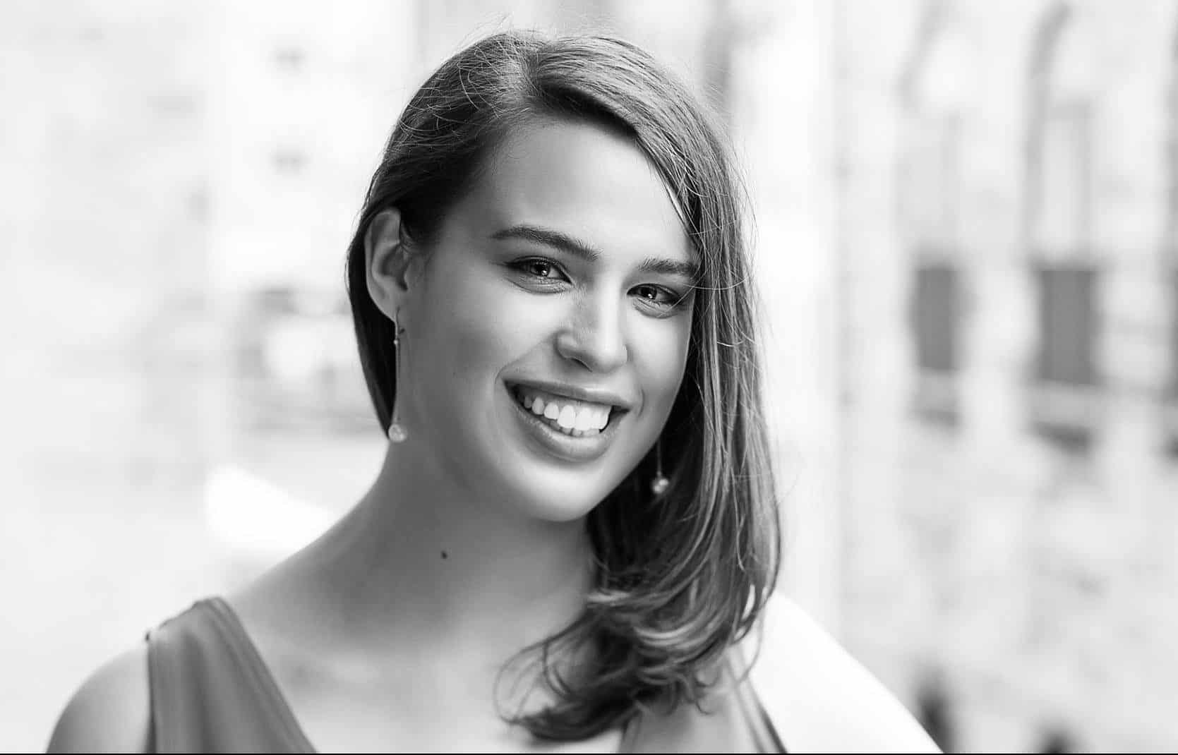 A young woman with straight, shoulder-length hair smiles at the camera. She is wearing a sleeveless top and drop earrings. The background is blurred and appears to be an outdoor urban setting. The photo is in black and white.