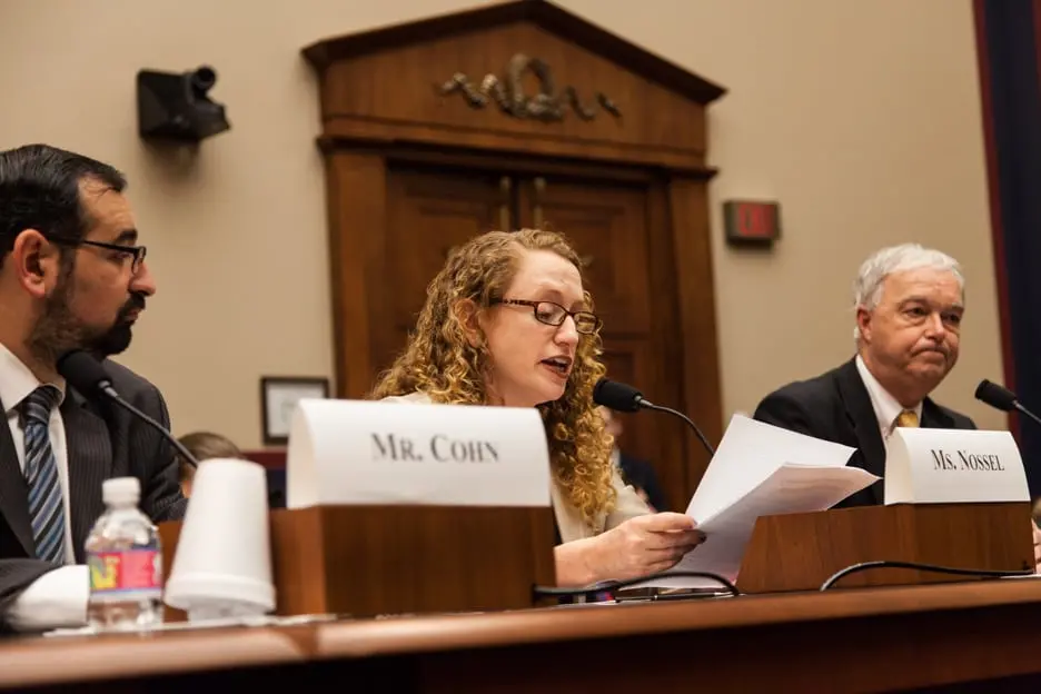 A woman with curly hair speaks into a microphone and reads from papers at a panel, seated between two men at a hearing. Name placards labeled “Mr. Cohn” and “Ms. Kinsel” are visible on the table.