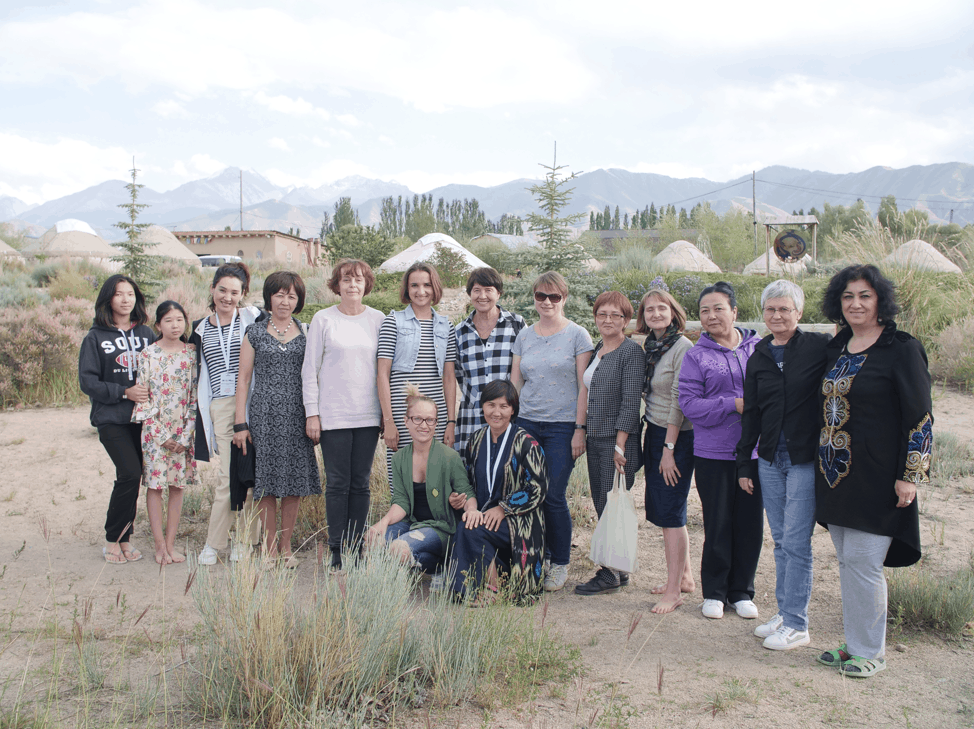 A group of people, mostly women, stand together outdoors in front of yurts and mountains. They are smiling and dressed in casual clothing, with some greenery and dry grass in the foreground.
