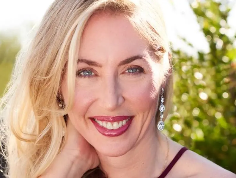 A woman with long blonde hair and blue eyes smiles outdoors. She wears dangling earrings and a sleeveless maroon top. Sunlight and greenery appear in the background.